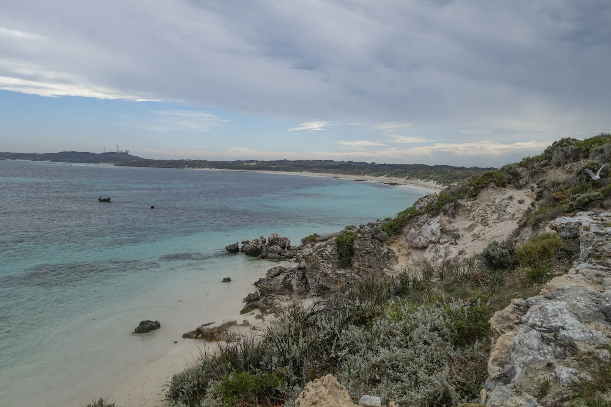 A turquoise bay with a sandy beach below a rocky coastal cliff